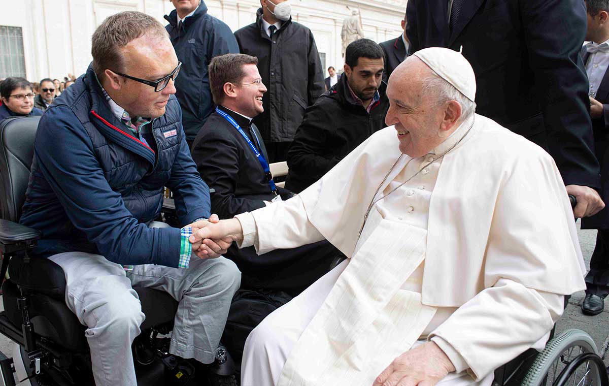 Man sitting in a power wheelchair shaking hands with Pope Francis, who is in a manual wheelchair.